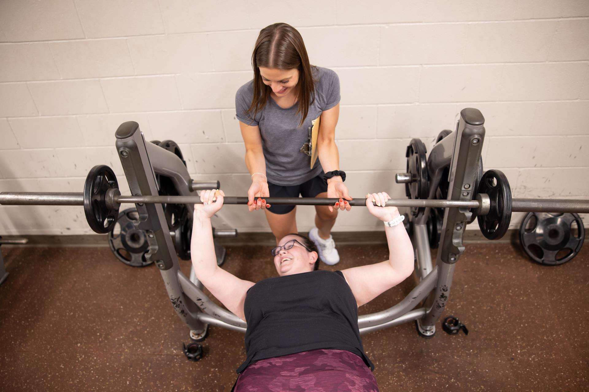 A woman lifting weights on a bench press while a personal trainer spots her