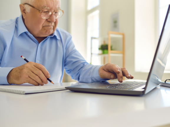 Older Man on Laptop While Writing in Notepad